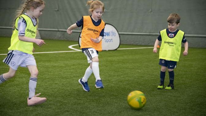 A child with an orange bib kicks a football on an indoor pitch, two other children are either side.