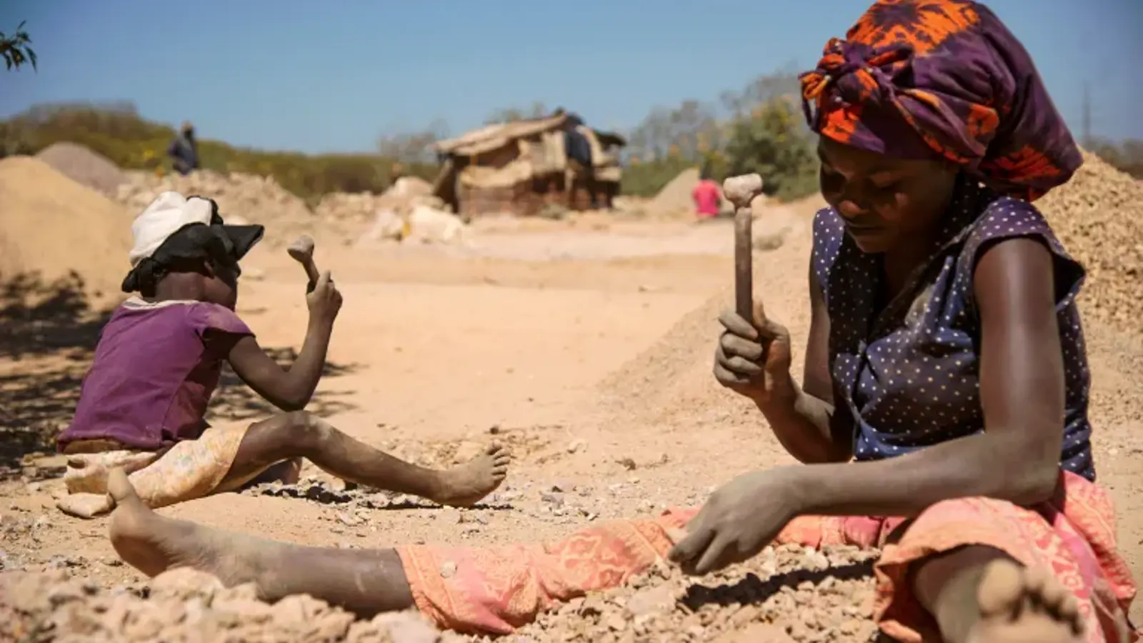 photograph of two people breaking cobalt from rocks at a sandy mine. One of the people is a child.