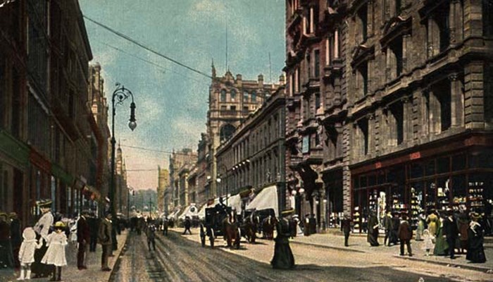 A photo of a busy Glasgow street with tall tenement style buildings on both sides, horse and carriage and lots of pedestrians.
