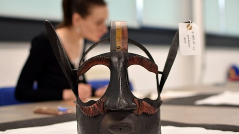 Photograph of a museum object with a blurred person in the background. The object is a mask made of metal designed to go over someone's head. It has ears, eyebrows and a moustache. The object label is attached to one of the ears and notes that it is a punishment mask.