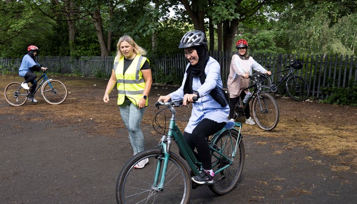 Three people learning to cycle with one other person in a high-vis vest helping them.