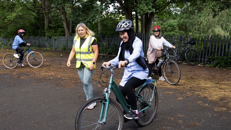 Three people learning to cycle with one other person in a high-vis vest helping them.