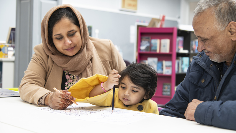 Two adults and a child taking part in a creative colouring activity at a library table, with bookshelves in the background.