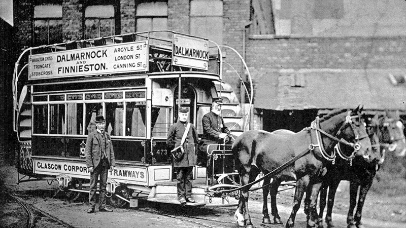 Black and white image of a Glasgow tram, two horses are pulling the tram, a man in a suit is standing in the foreground and two men are on the tramnd 