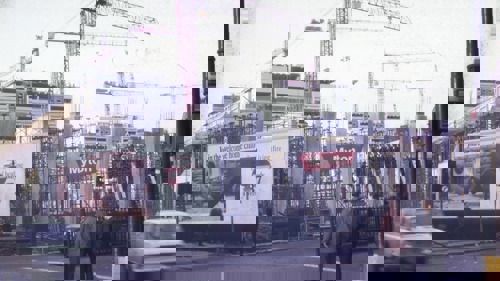 A colour image of the Anderston Centre under construction, in 1970. There are two men waiting to cross the road in the foreground, and a white car is driving in the direction of the camera. Over the road there is a building site, with the name 'Myton' in red lettering affixed to the fence. There is a billboard in the mid-ground, and in the background there are several red cranes.