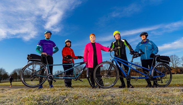 Five people standing holding tandem bikes on grass with blue sky and few clouds above