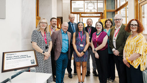 A group of ten people gathered for a photo in a historic library building with stained glass details. They are standing next to a plaque that reads Elderpark Library and Community Hub
