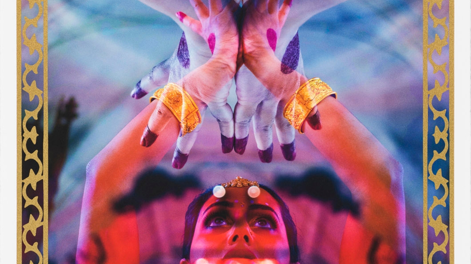 A vivid, symmetrical artwork shows a central figure with upraised hands adorned in gold bracelets. The hands are painted in bold red and purple tones, and the face below them glows with red light, eyes lifted upward. The composition has a mirrored, layered effect that gives it a surreal, almost spiritual intensity. A decorative gold border frames the image, and small text at the bottom includes edition information and artist names.