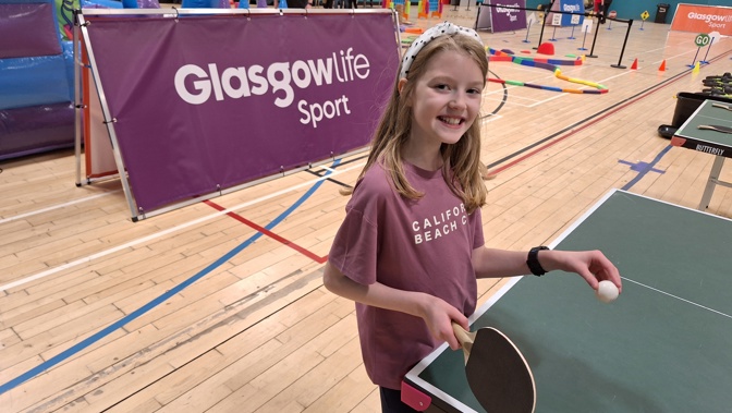 A child standing at a table tennis table with a racket and ball in front of a Glasgow Life Sport sign.
