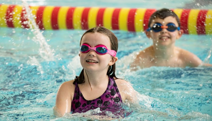 Two young children wearing goggles and smiling while they splash in swim in a swimming pool
