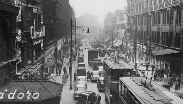 A black and white photo of a busy street, with tall buildings on both sides of the road and includes many pedestrians, cars and trams traveling.