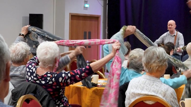 A musician performing in front of an older crowd at a dementia-friendly concert, some of the crowd are holding scarves above their heads.
