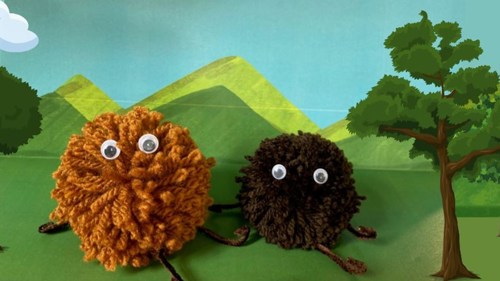Two pom pom haggis characters with googly eyes in front of a painted background showing mountains and trees