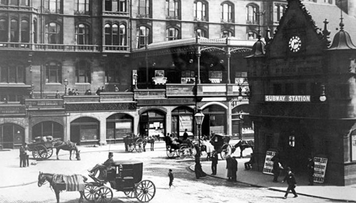 A black and white photo of a square in Glasgow, with cobbled road, horse and carraige, people and entrance to a subway, with large buildings with many windows in the background.
