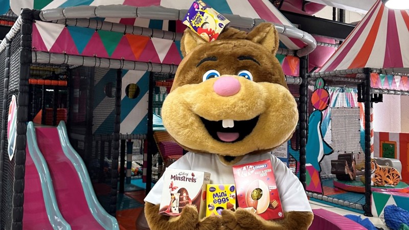 A squirrel mascot is holding chocolate easter eggs, in the background is a children's soft play area