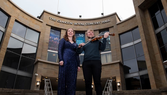 One person in a sparkly blue dress and another next to them holding a violin and bow standing on the stairs in front of Glasgow's Royal Concert Hall