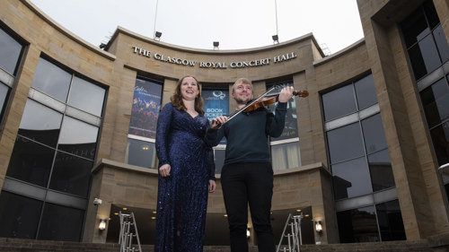 One person in a sparkly blue dress and another next to them holding a violin and bow standing on the stairs in front of Glasgow's Royal Concert Hall