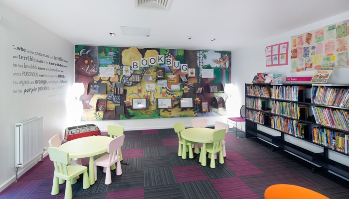Children's area with lime green and baby pink tables and chairs. There is children's artwork on the walls and a sign that reads 'Bookbug'.