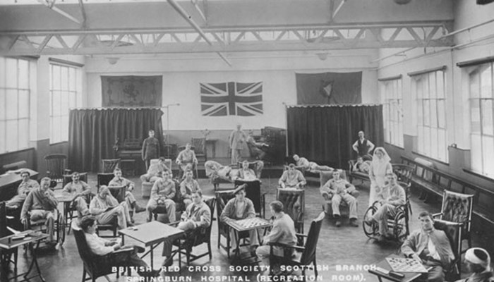 A black and white photo of several people in a recreational ward sitting in chairs playing games such as checkers, some have bandages from injuries sustained. There is a union jack flag on the wall.