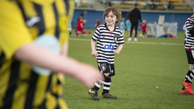A child, wearing a black and white football strip, smiles widely while on a grassy football pitch.
