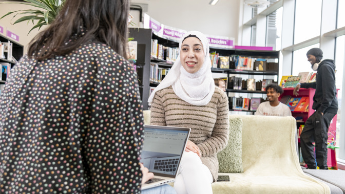 Person wearing a light-colored headscarf and striped sweater sits on a sofa in a bright library, smiling while using a laptop. Another person sits nearby with a laptop, and bookshelves and other people browsing books are visible in the background.