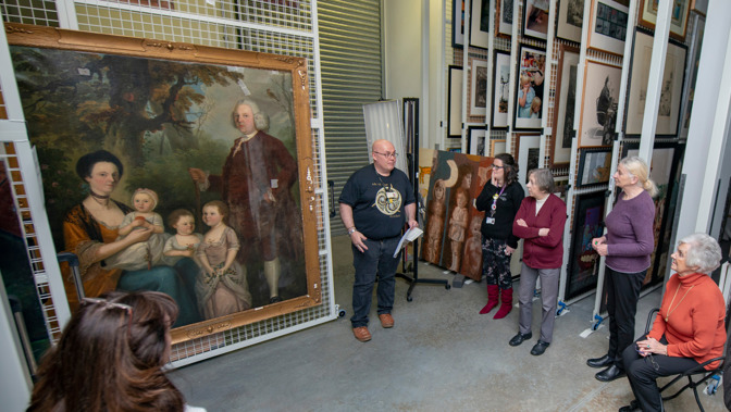 A group of people stand and sit in a museum storage area listening to a guide who is speaking and holding papers beside a large framed historical family portrait; racks of framed artworks are visible behind them.