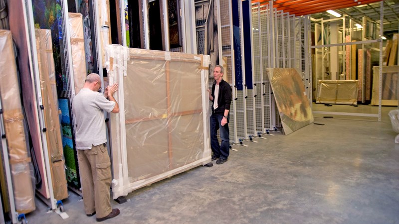 Two technicians are preparing to move a large painting wrapped in bubble wrap at Glasgow Museums Resource Centre