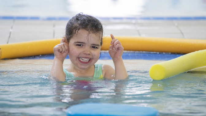 A young girl grins happily while playing in a swimming pool, surrounded by floating pool noodles and water toys.