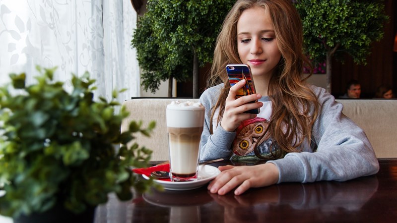 Young adult sit at a table looking at their phone. There is greenery around them and they have a delicious looking creamy coffee in front of them