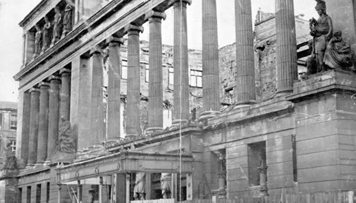 A black and white photo of a building destroyed by fire. Pillars and statue remain, the building is fenced off and people walk the pavement outside of it and cars are parked on the road.