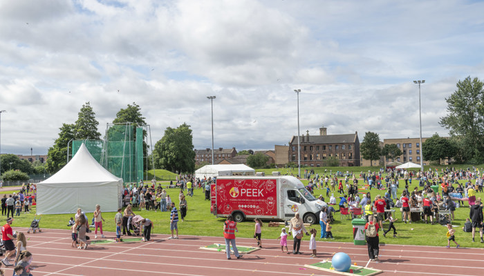 Large outdoor community play day with crowds, activity tents and a PEEK van on a sports field and running track.