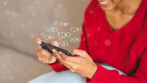 A person of colour holds a smart phone with social media icon graphics floating out of it.