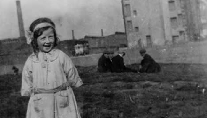 A black and white photo of a young child smiling and standing on a patch of waste ground. In the background a group of people are gambling. 