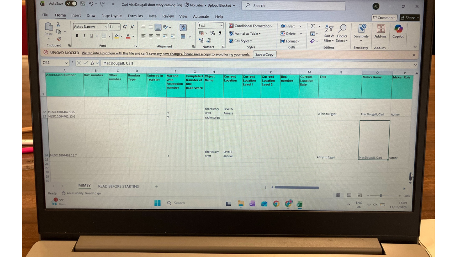 A laptop screen displaying an Excel spreadsheet titled “Carl MacDougall short story cataloguing,” showing rows of cataloguing data for drafts and a radio script of “A Trip to Egypt,” along with a warning banner stating that the file cannot be saved.