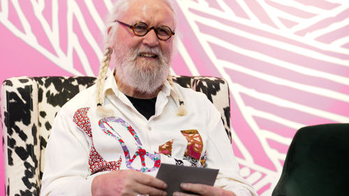 Billy Connolly sitting in a chair with a pink background looking and smiling to the front