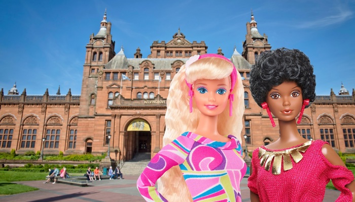 Two fashion dolls are posed in front of the frontage of Kelvingrove Art Gallery and Museum, a large, ornate, red sandstone building on a sunny day. The doll on the left has long blonde hair and wears a bright patterned pink, purple, and green dress with pink accessories. The doll on the right has short curly black hair and wears a red dress with gold details. Behind them, the museum’s red sandstone frontage, towers, and arched entrance are visible, with people sitting and walking in the foreground. 