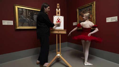 A young female artist sketches a young ballet dancer dressed in a beautiful traditional handmade ballet outfit in front of Degas artworks on a red wall in the Burrell Collection 
