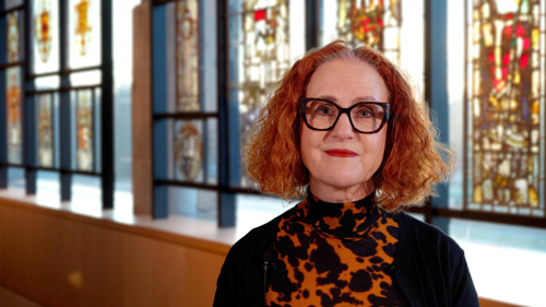 A person with shoulder length red hair, wearing black rimmed glasses and a brown and black polo neck top is standing in a museum gallery displaying stained glass windows