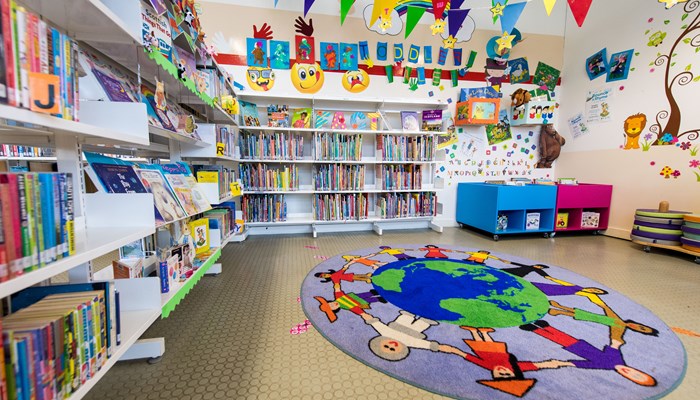 A very colourful children's area in Barmulloch Library with bookshelves, bunting, posters and a round rug of the world with funny faces surrounding it