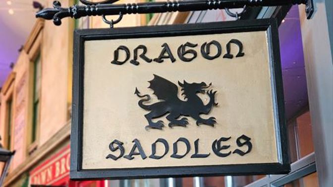 Hanging, carved wooden sign for "DRAGON SADDLES" in a historic-looking street display.