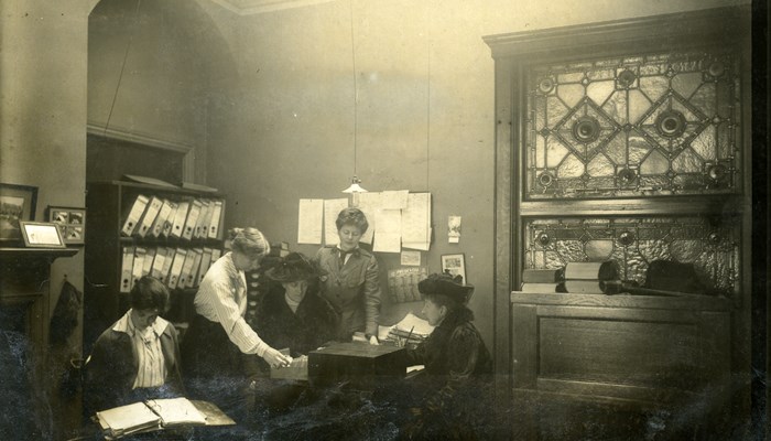 Black and white photo of 5 people in The Scottish Women's Hospital office, one of which is signing papers. Papers are hanging on the wall and a filing cabinet is behind them.
