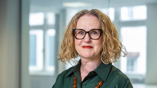 Headshot of a smiling person in an office. They are wearing glasses and a large colourful necklace.