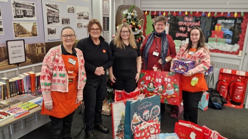 A group of five people surrounded by Christmas gift bags in a community centre. Some of them are wearing orange aprons with B&Q written on them and Christmas jumpers
