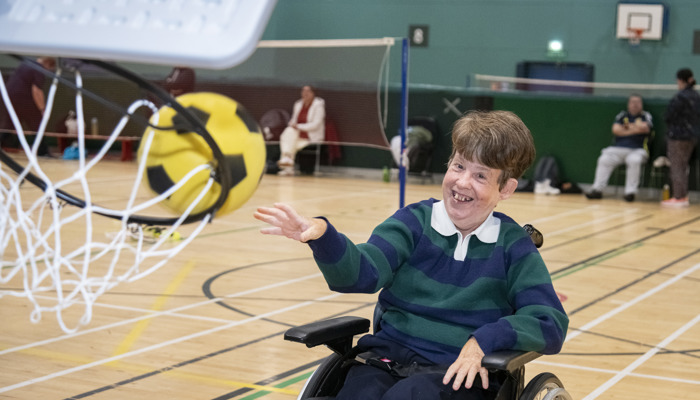 A person in a wheelchair smiles while throwing a large yellow and black ball near a basketball hoop.