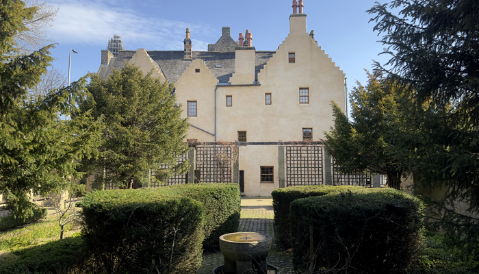 Exterior view of a historic cream-coloured building with stepped gables and multiple chimneys, seen from a formal garden with neatly trimmed hedges and a central stone feature in the foreground. The building has small rectangular windows and a slate roof, with tall trees framing both sides and a clear blue sky overhead.