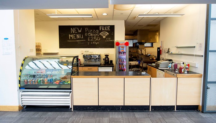 A food counter with a glass display case containing juice cans and crisps to the left. There is a chalkboard that says 'New Menu' in the background.