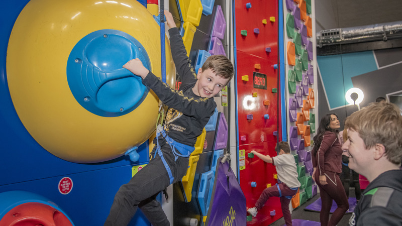 Child in a safety harness climbing an indoor climbing wall in a brightly coloured activity centre, with other climbers and spectators nearby.