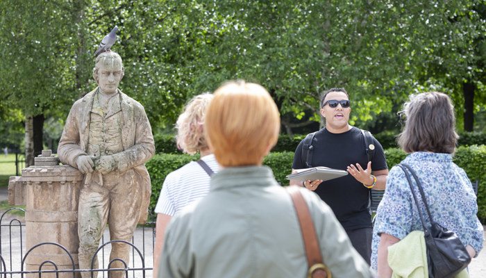 Guide speaking to a small group beside a stone statue with a pigeon perched on its head in a leafy park.
