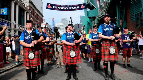 Bagpipers in tartan kilts perform outside Fenway Park beneath a welcome banner as crowds gather along the street.