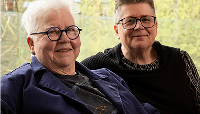 Two women with grey and dark hair are wearing dark jackets while sitting down